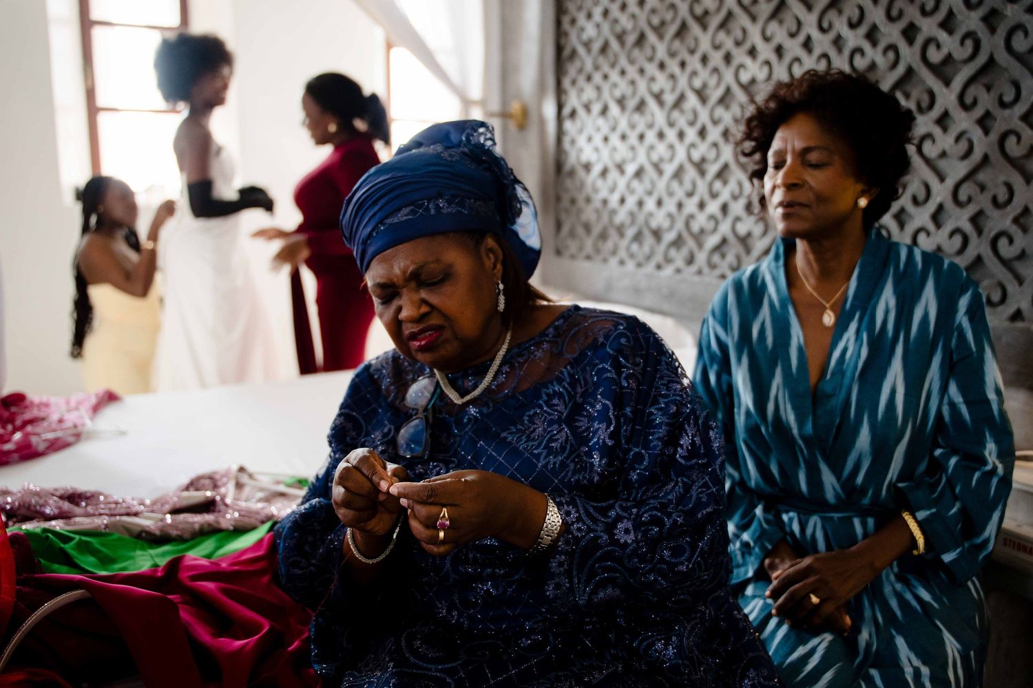 People gather in a room while someone in a blue head wrap and dress sits in the foreground.