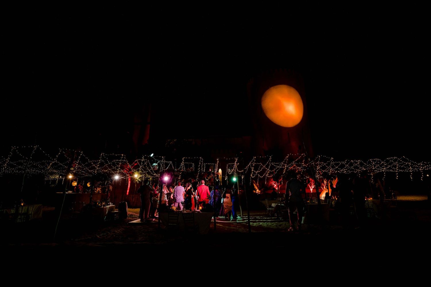 A large orange moon rises over a nighttime concert stage with colorful lights and silhouetted crowd.