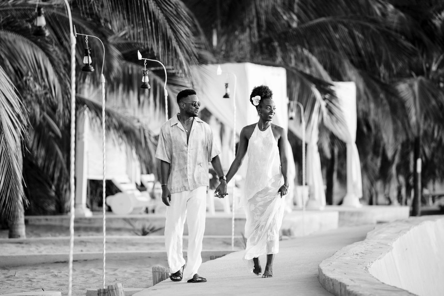 A couple in white outfits walking together on a palm-lined path in a black and white photograph.
