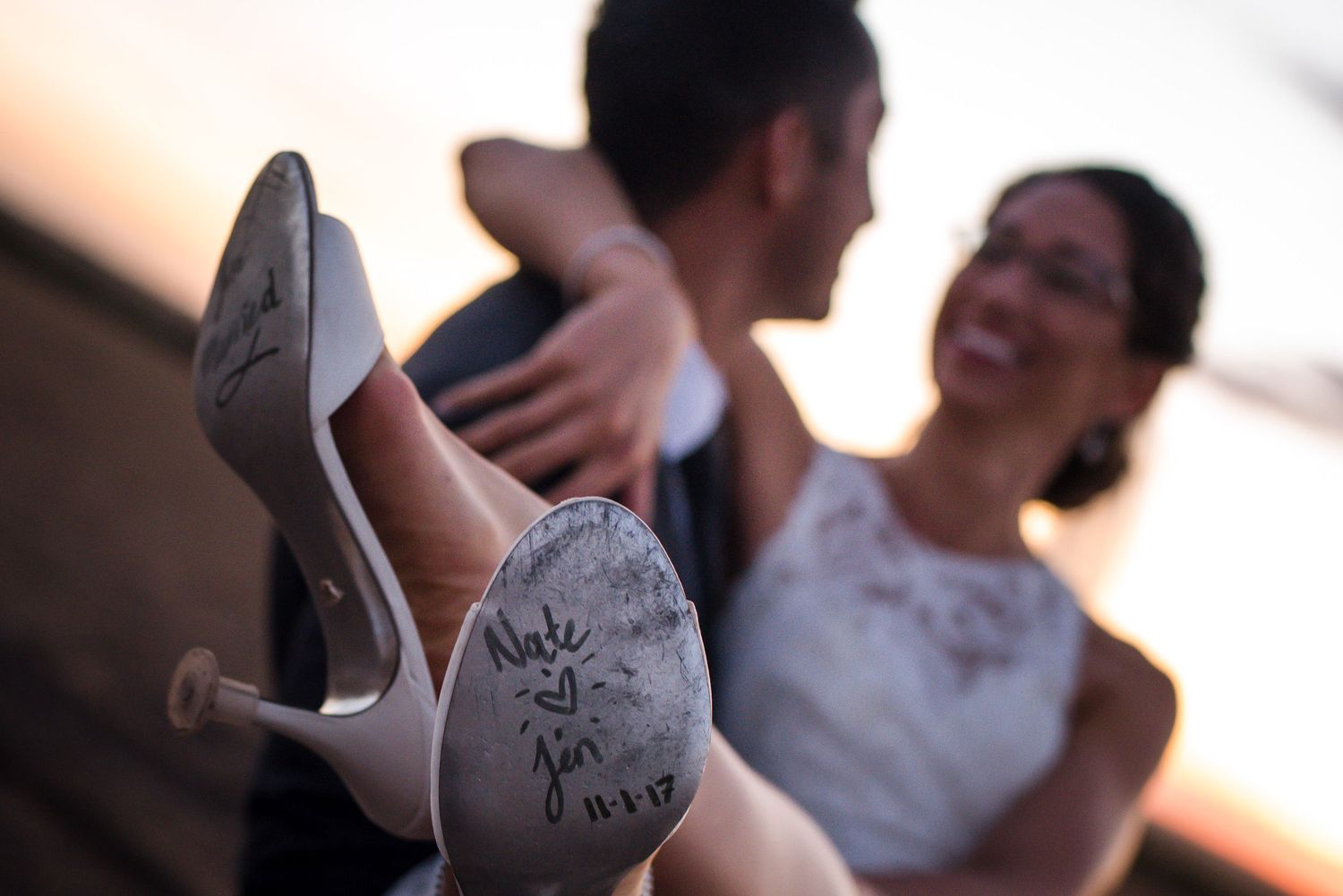 Close-up of bride's heel with wedding date written on sole, happy couple embracing in soft focus background.