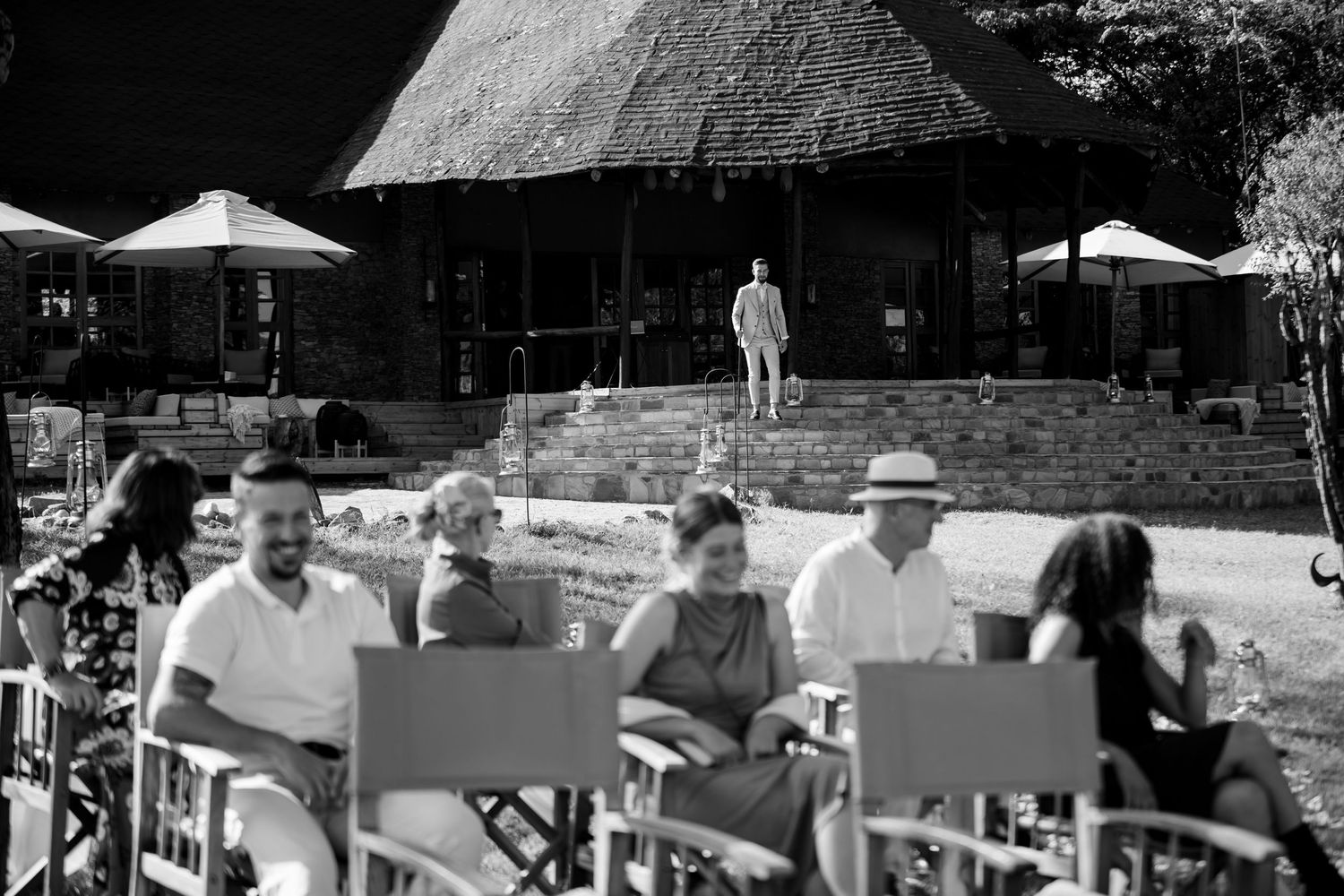Black and white photo of guests relaxing in chairs at an outdoor safari lodge with thatched roof building.