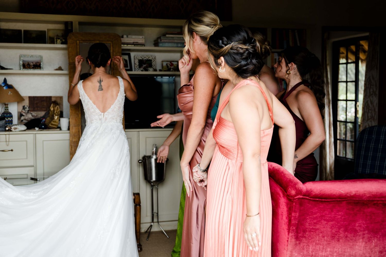 Bridesmaids in peach colored dresses help adjust the bride's wedding gown in a sunlit room.