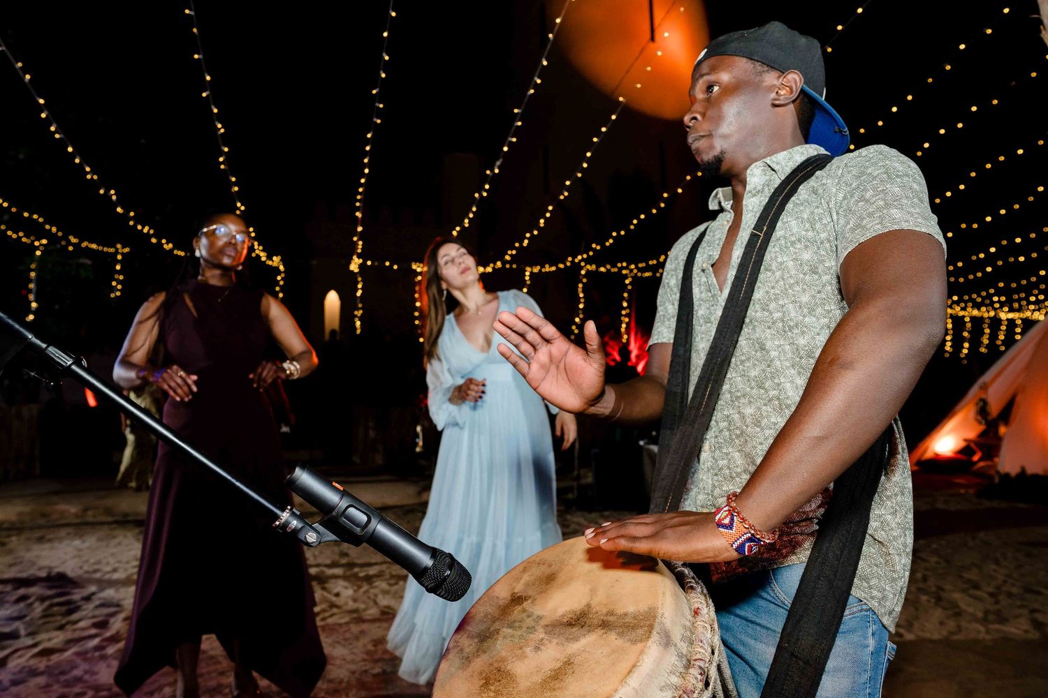Musicians perform at a nighttime outdoor celebration with string lights overhead and a drum in the foreground.