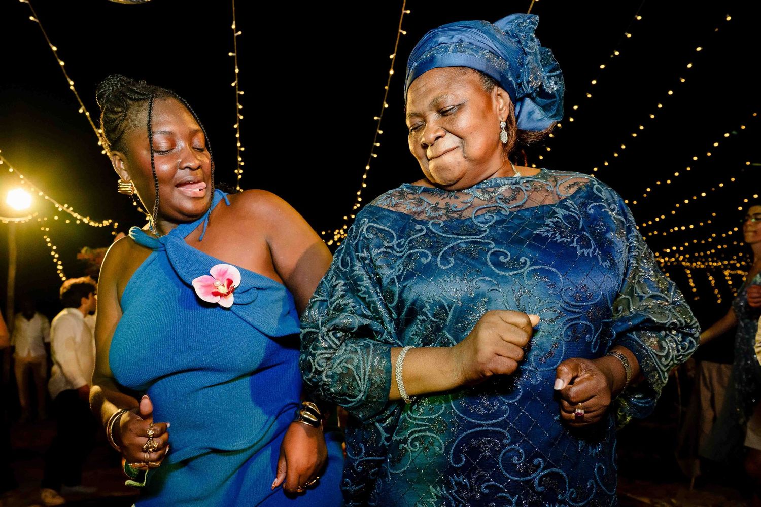 Two women dancing joyfully at a nighttime celebration, one in blue formal dress and another in embroidered traditional attire.