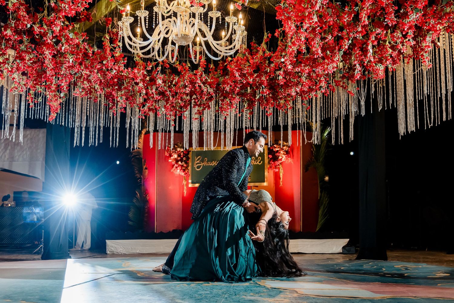 Dancers performing under an ornate ceiling decorated with red flowers and crystal accents.