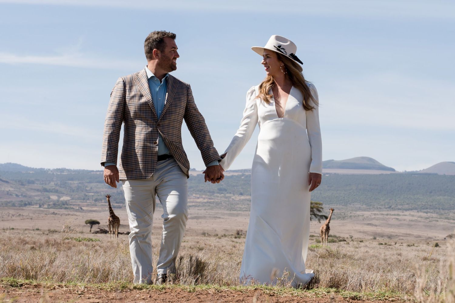 A couple holds hands while walking through the African wilderness with mountains in the distance.