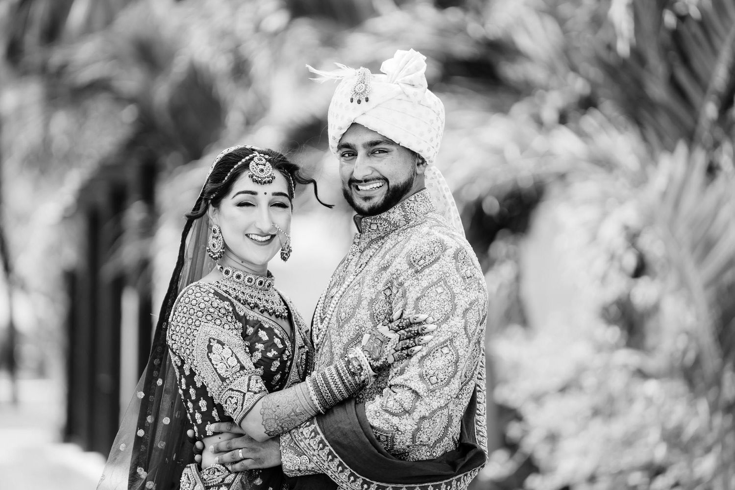 Black and white photo of a couple in traditional Indian wedding attire smiling together outdoors.