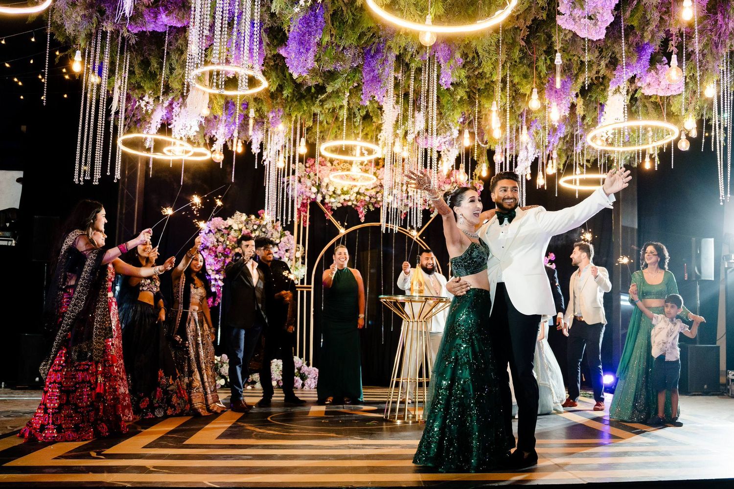 Wedding reception guests dancing and celebrating under elegant hanging lights and decorative floral arrangements at an evening party.