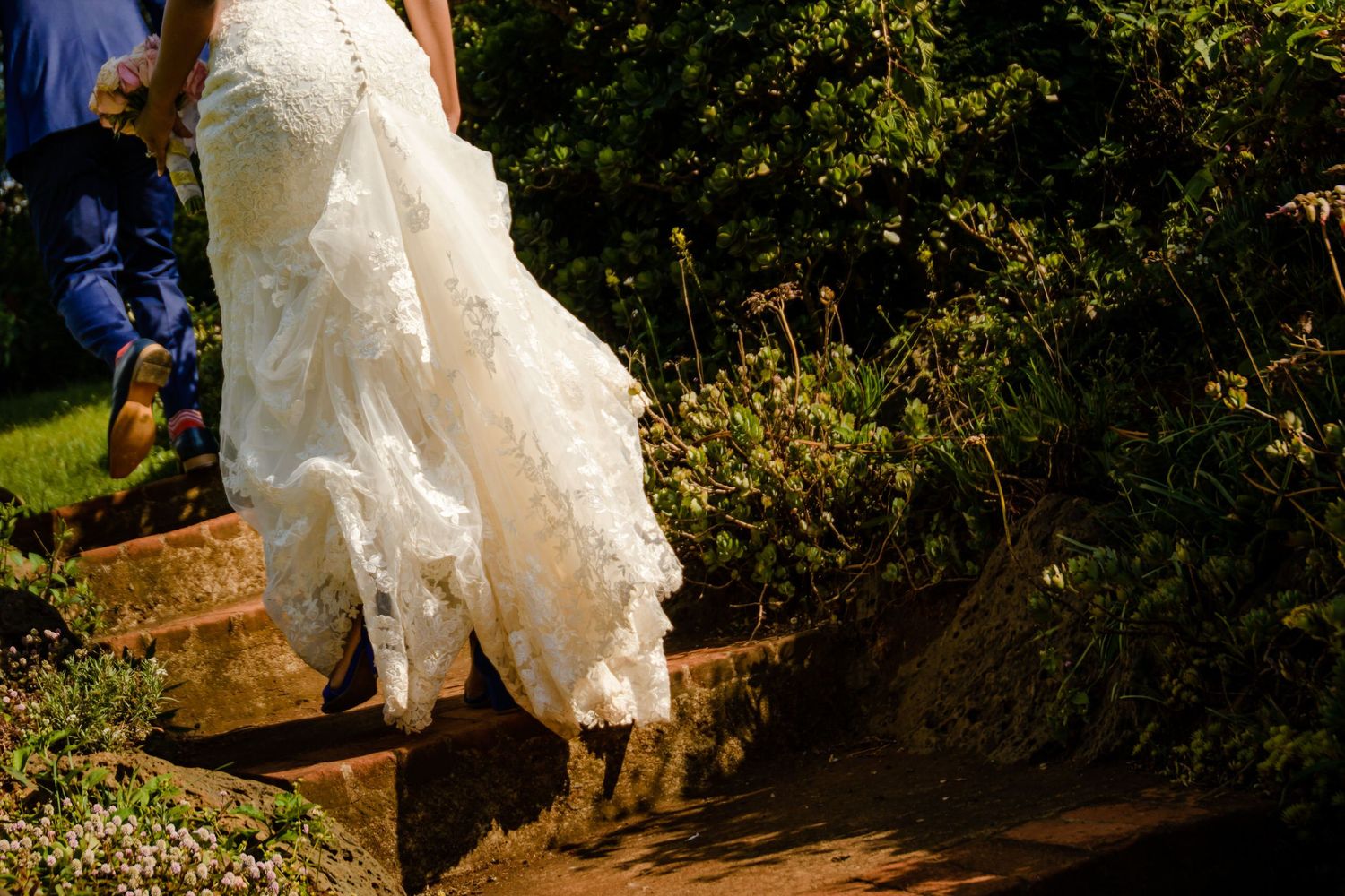 Close-up of a white wedding dress hem and boots on rustic wooden steps in a garden setting.