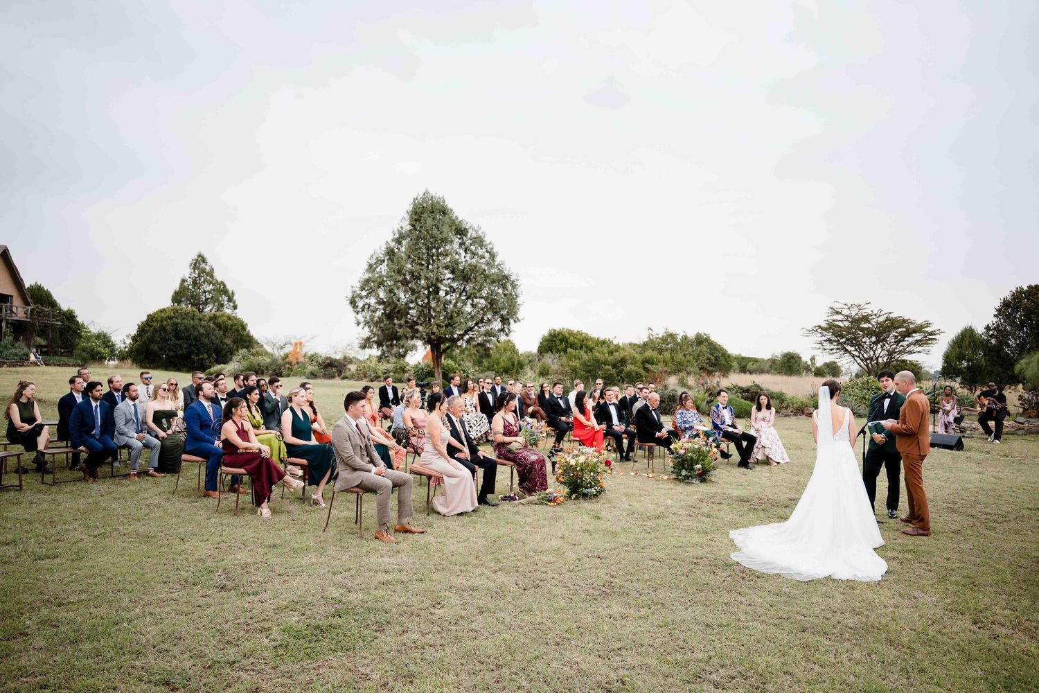 An outdoor wedding ceremony takes place on a grassy field with guests seated in rows and a couple standing at the front.