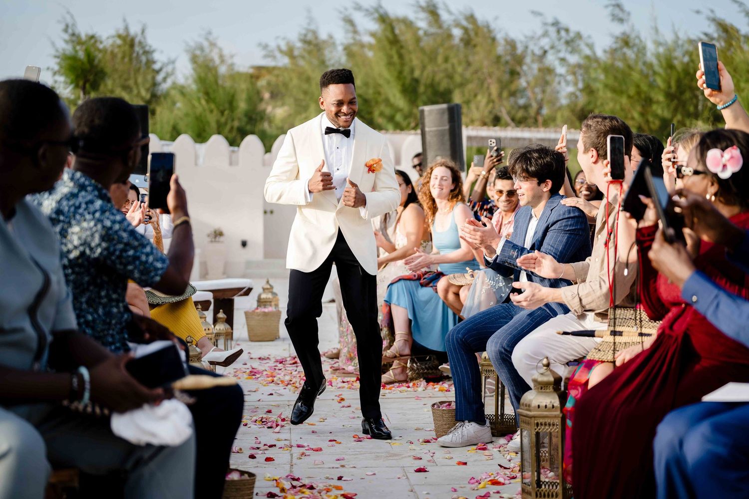 Wedding guests throw red rose petals during an outdoor celebration ceremony at a sunny garden venue.