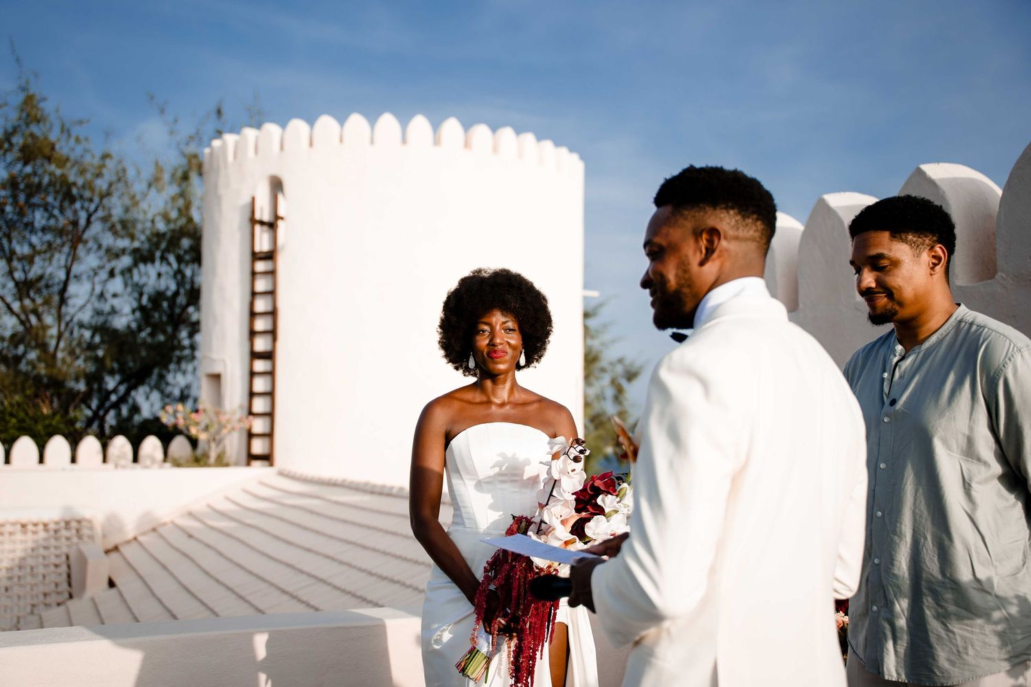 A wedding couple stands together with a guest at a venue with a white cylindrical tower and blue sky in the background.