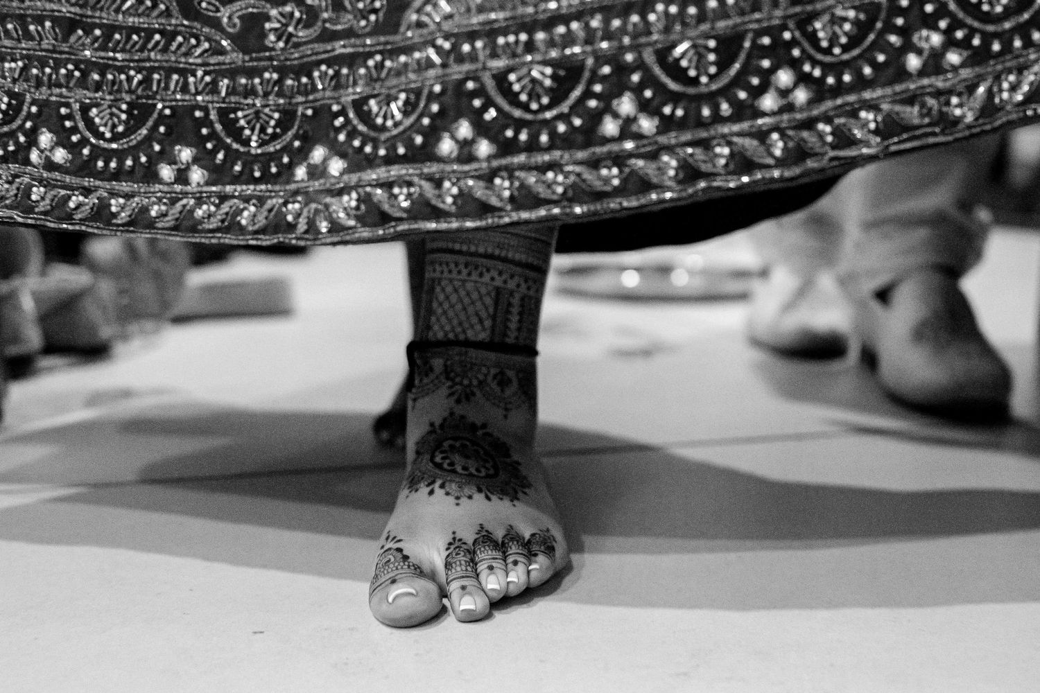Black and white close-up of decorated feet with traditional henna and jewelry beneath an ornate garment hem.