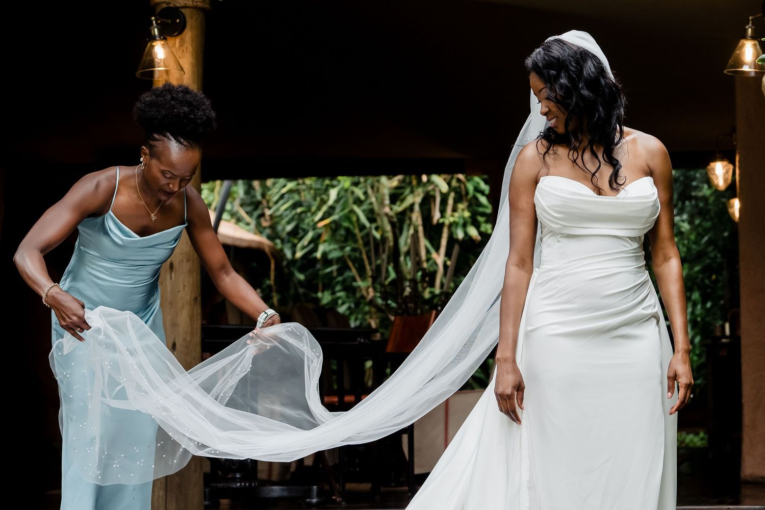 Two people adjust the flowing train of a white wedding dress outdoors.