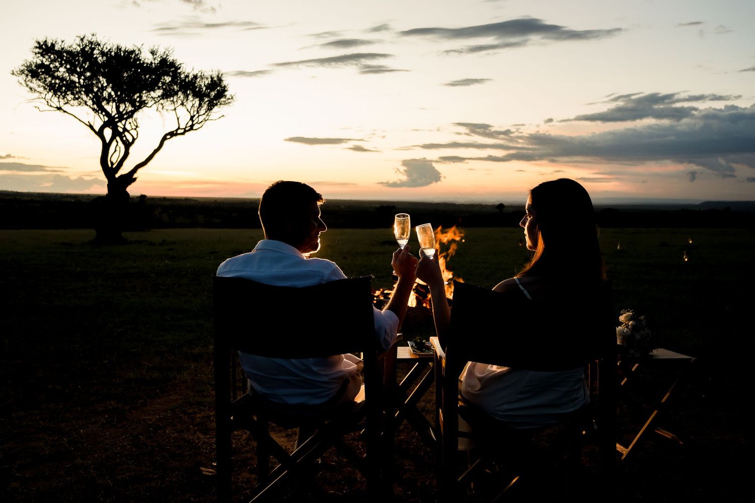 Couple toasting champagne at sunset on African safari, silhouetted against dramatic sky near acacia tree.