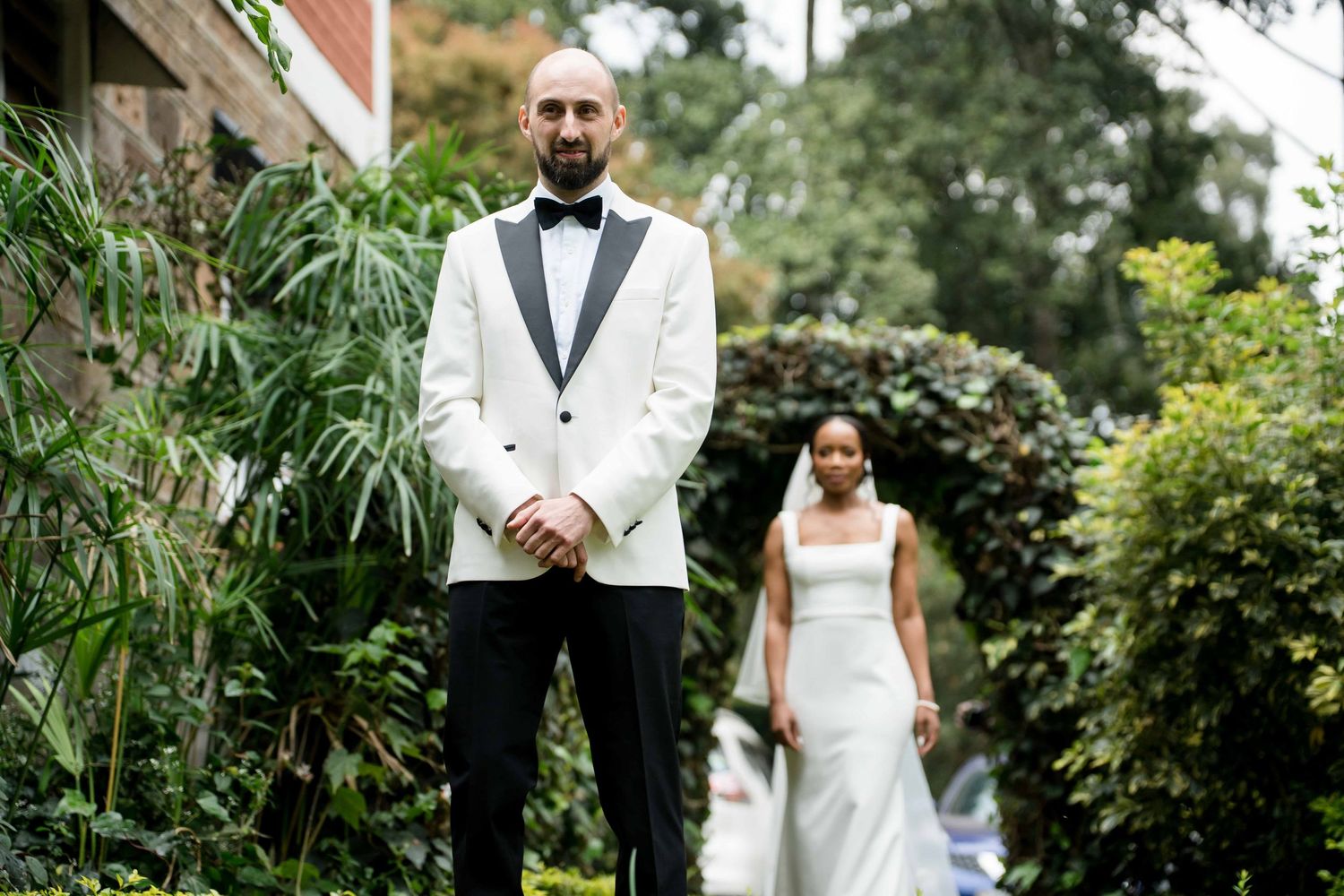 A couple in wedding attire stands together in a lush garden setting during their outdoor wedding ceremony.