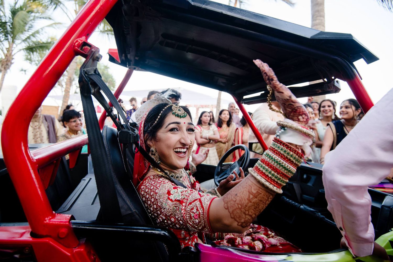 Joyful bride in traditional red Indian attire celebrates from inside a red car during wedding festivities.