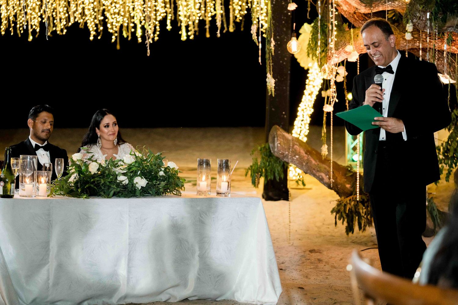 A luxurious wedding reception features elegant table settings and hanging floral decorations under warm lighting.