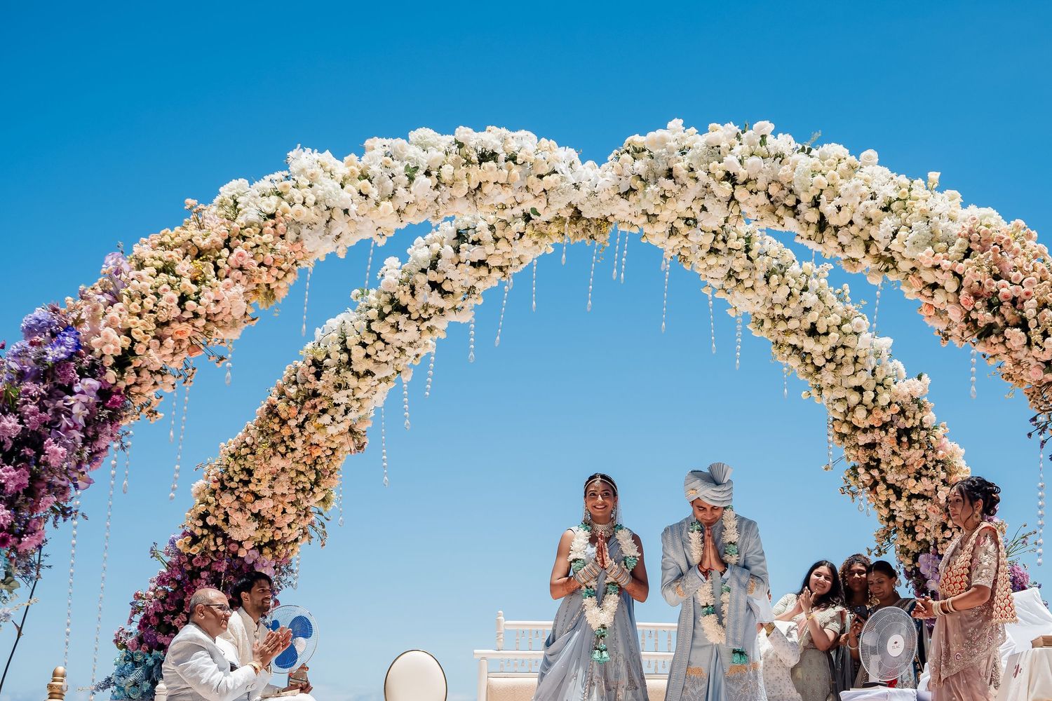 Outdoor wedding ceremony setup with elegant floral arches against bright blue sky.