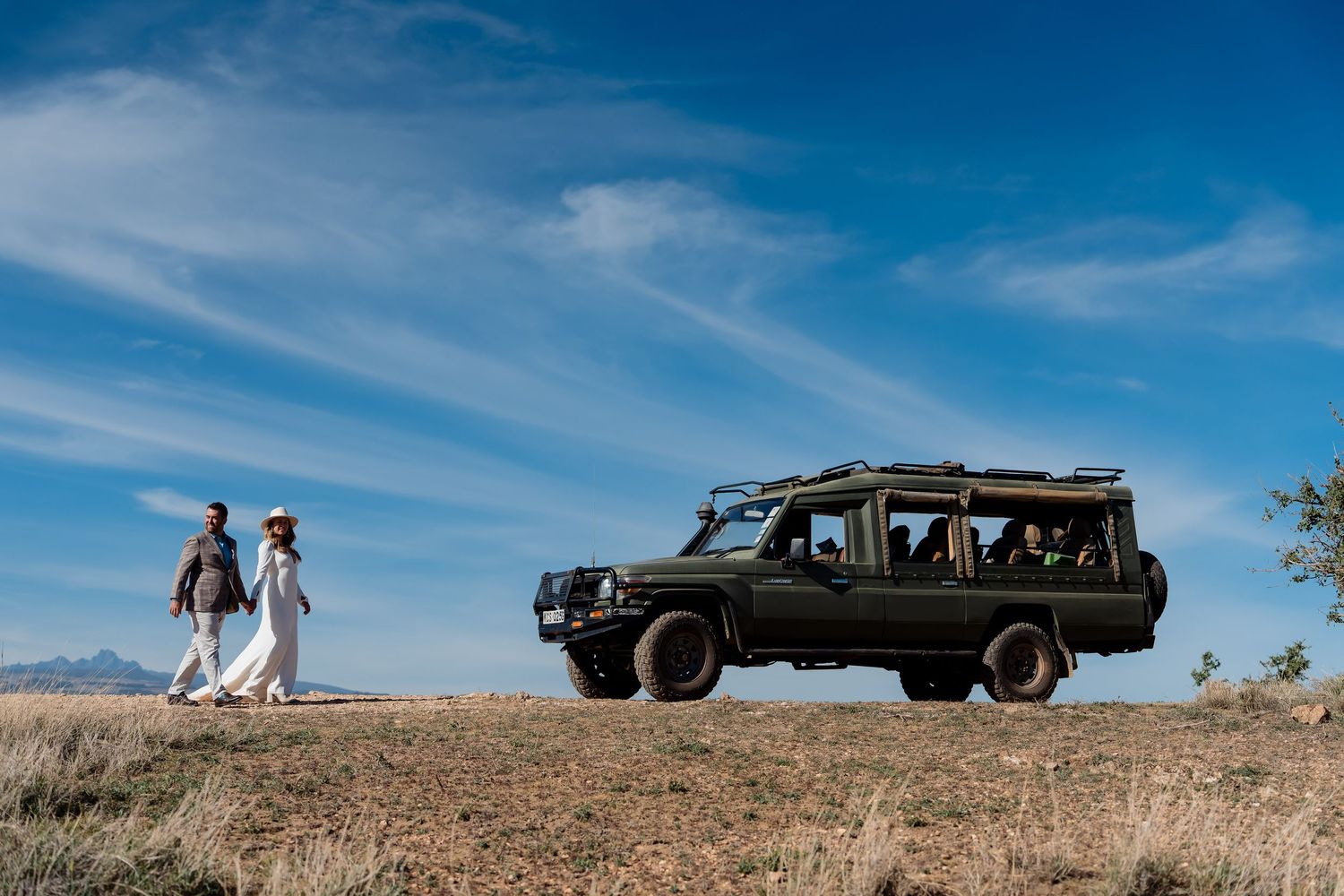 A black safari vehicle sits on a hill as two people walk in the distance under blue skies.