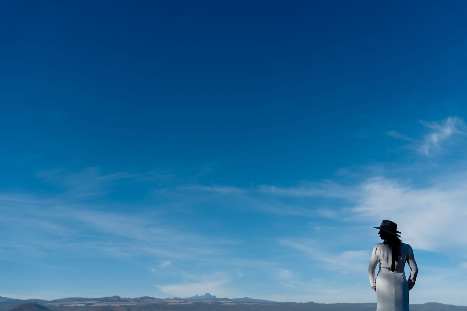 A silhouetted figure stands against a vibrant blue sky and mountainous landscape in white attire.