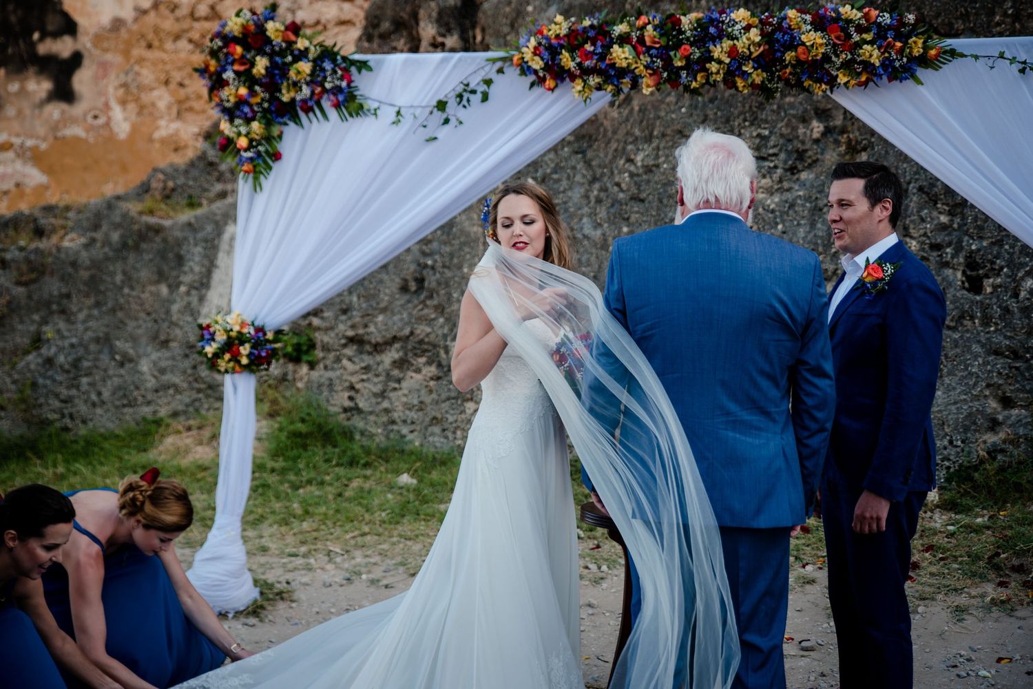A wedding ceremony takes place outdoors with decorative white fabric draping in the background.