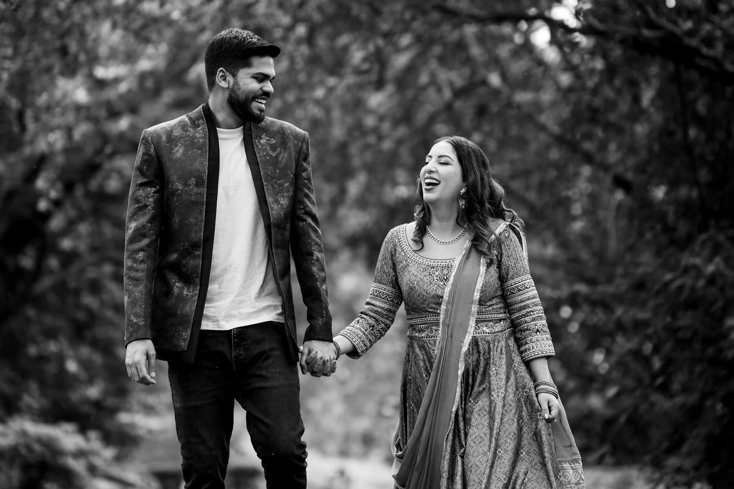 A couple laughs together while walking hand in hand through a wooded area in black and white.