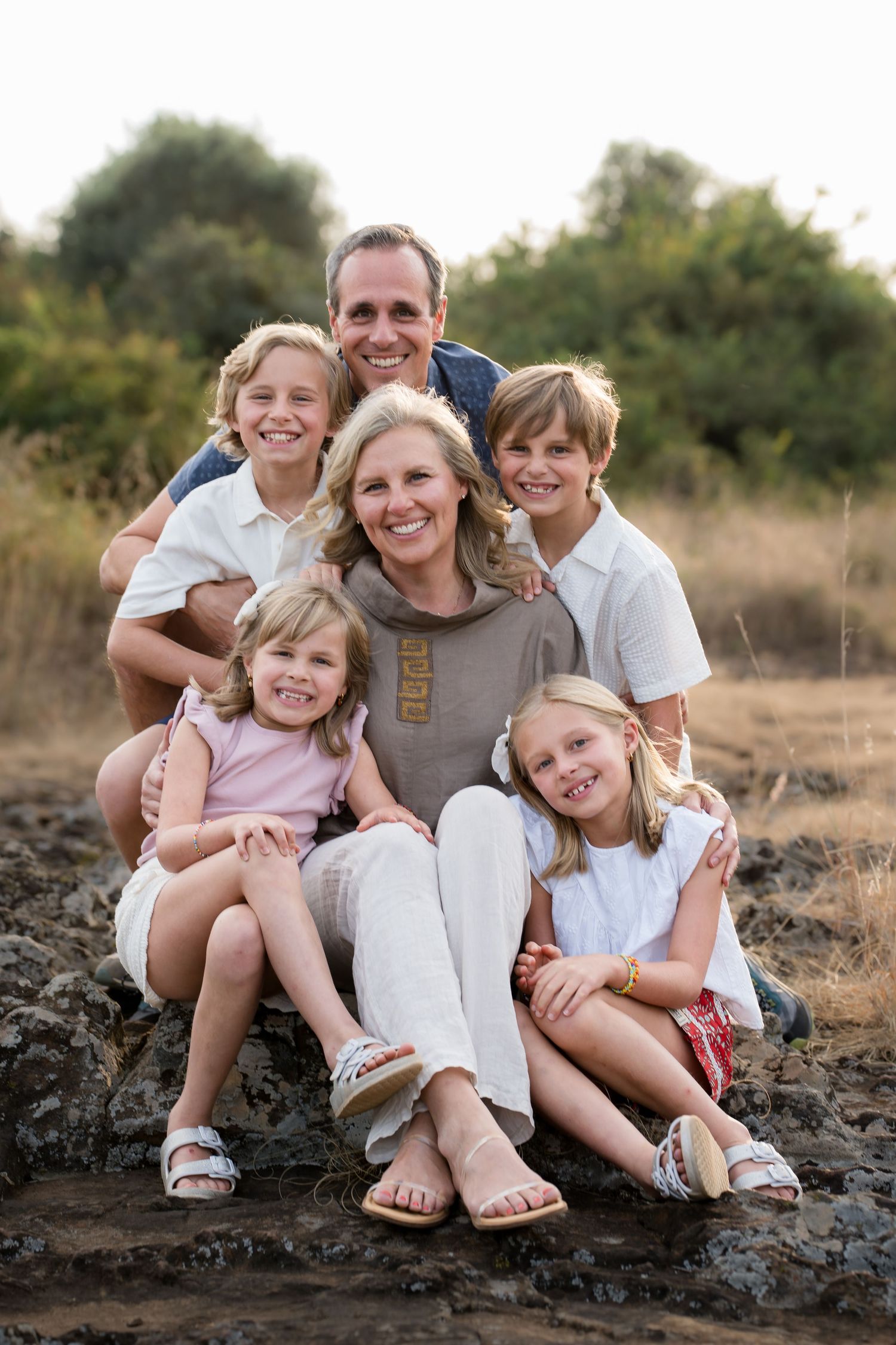 A happy family of six sits together outdoors on rocky terrain during golden hour in casual attire.