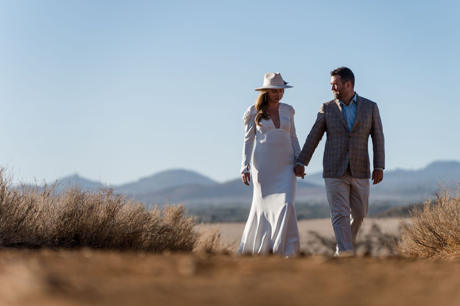 Couple in wedding attire walking through desert brush with mountains in background.