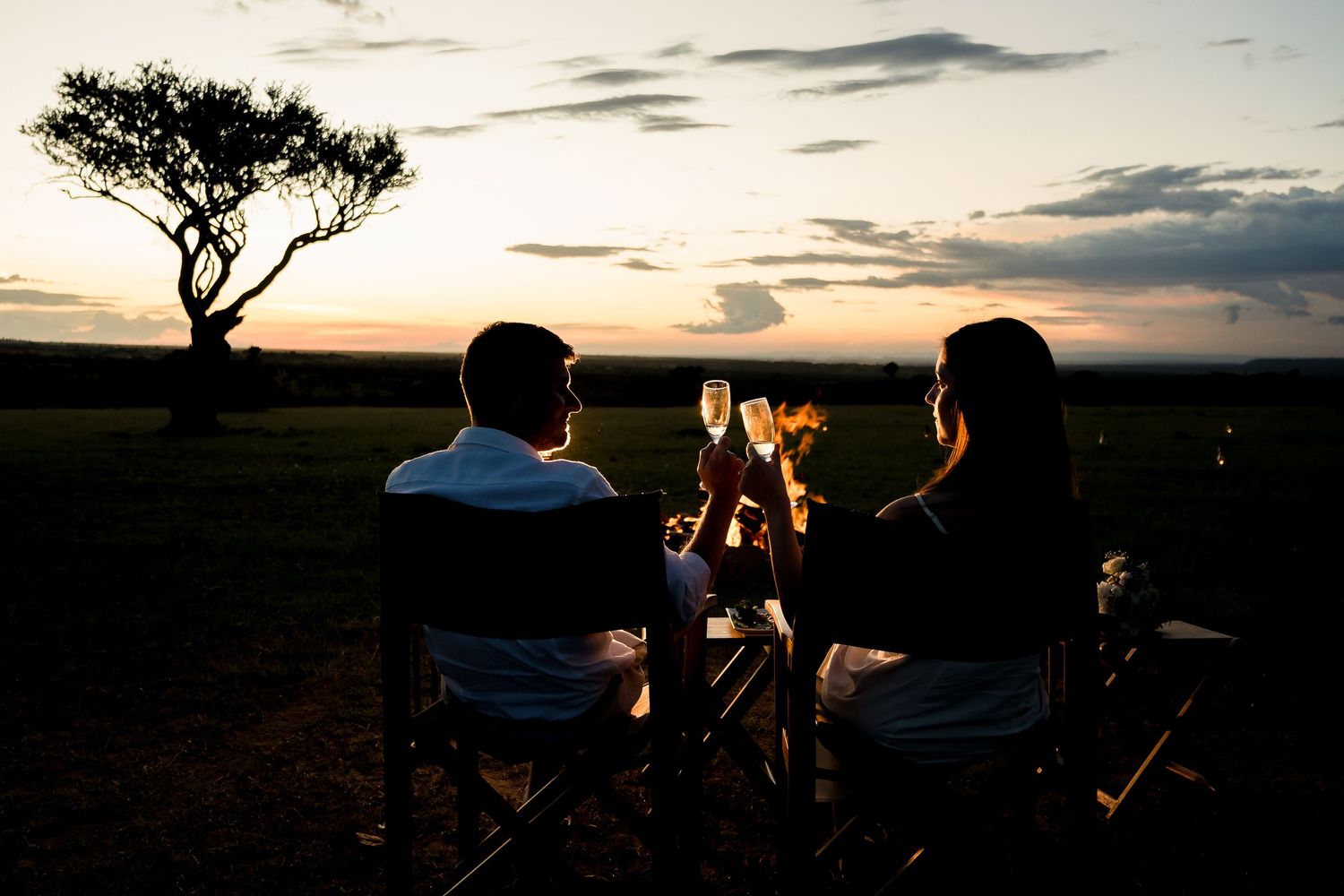 A couple enjoys drinks by a campfire under an acacia tree at dusk.