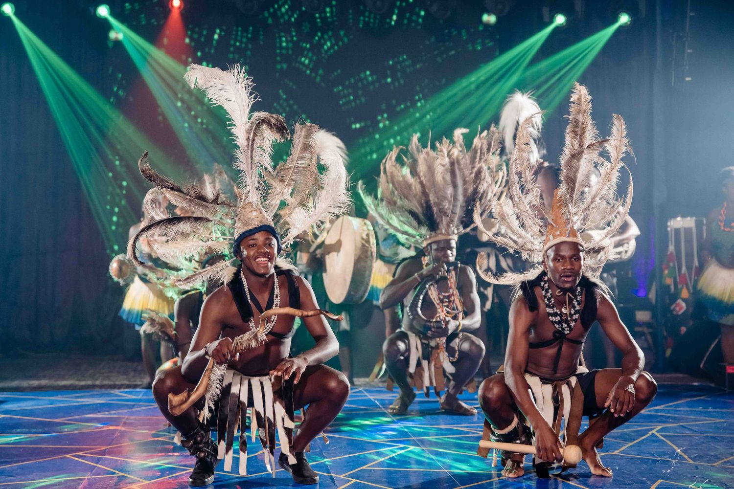 Traditional dancers perform in elaborate feathered costumes under vibrant green stage lighting.