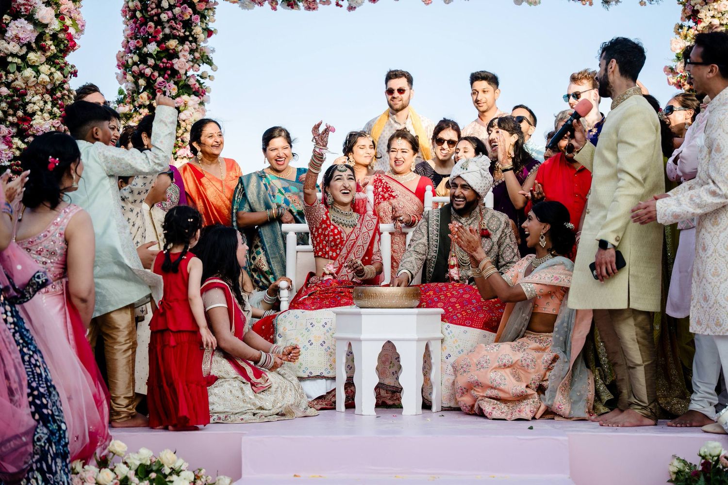 Wedding ceremony taking place under floral arch with guests gathered around traditional seating arrangement.