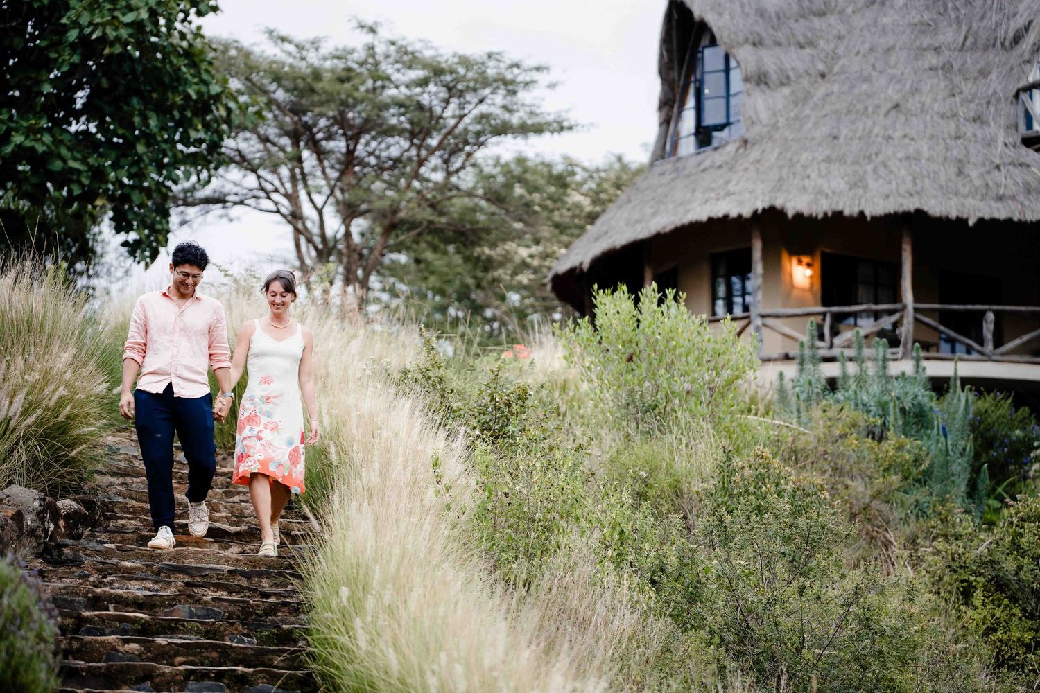 A couple walks through tall grass near a rustic thatched-roof building on a sunny day.