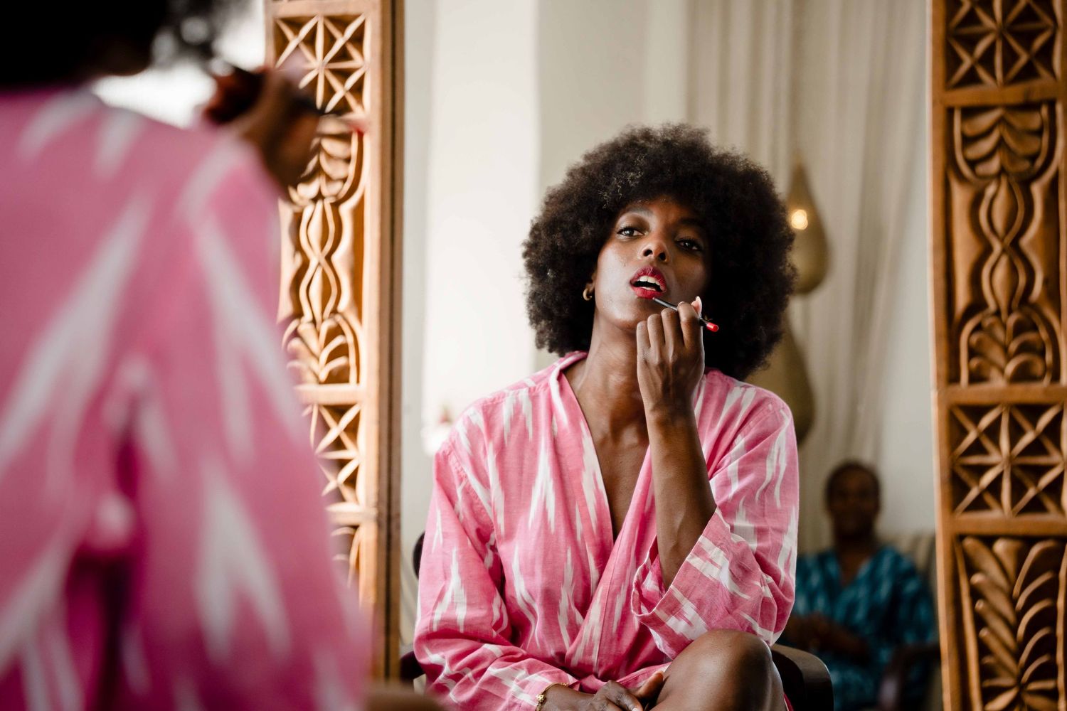 Woman in pink robe sits by ornate window screen with natural light streaming in.