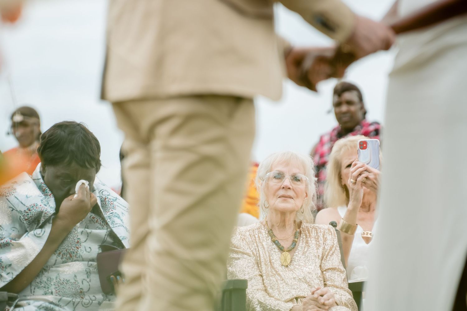 Elderly woman seated outdoors at a wedding ceremony, surrounded by guests holding hands and taking photos.