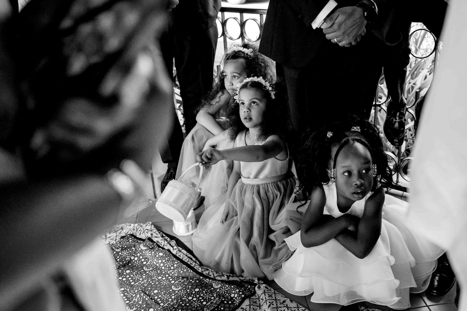 Black and white candid photograph of young flower girls in white dresses sitting on the ground during a wedding ceremony.