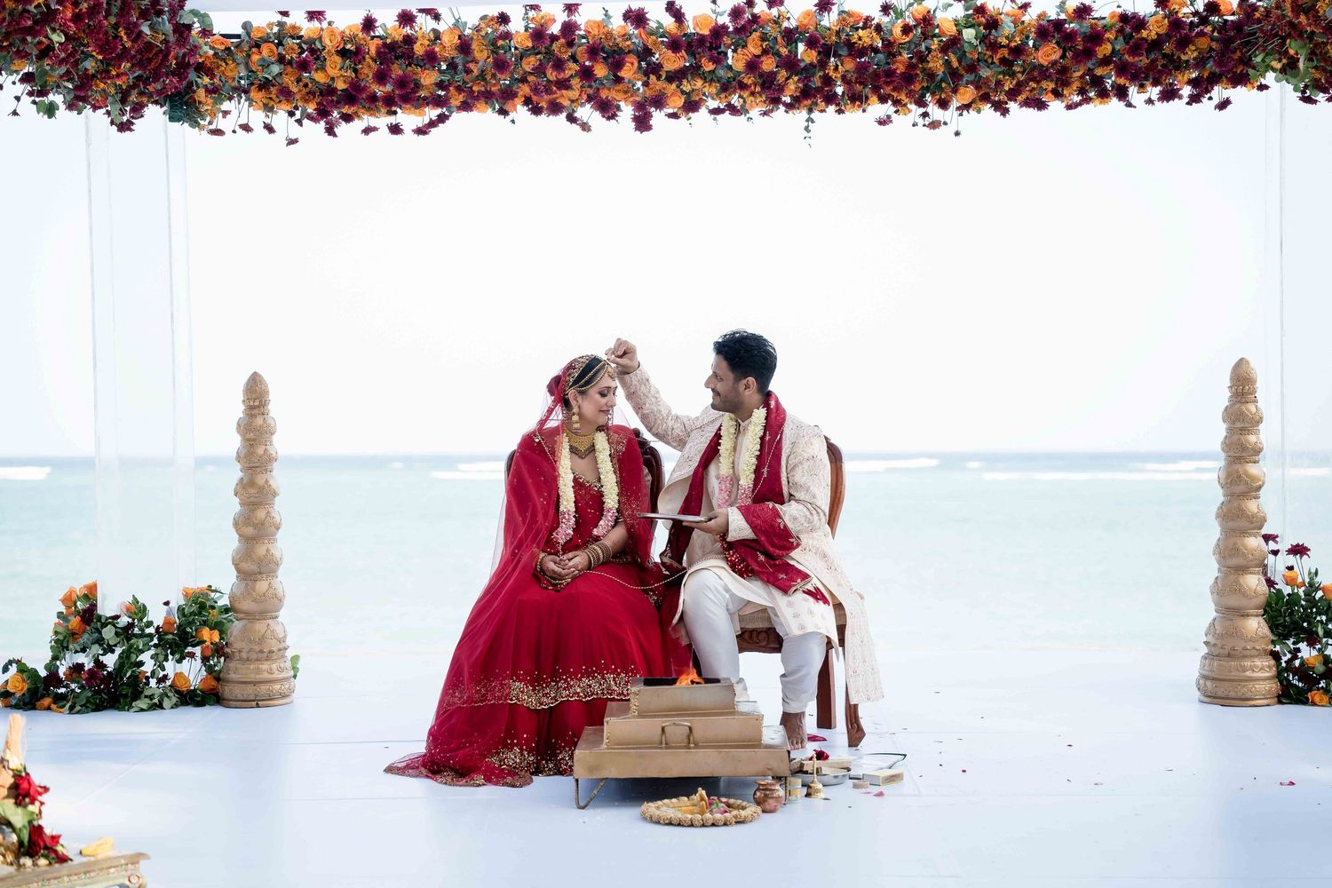 A traditional Indian wedding ceremony with red and gold decor and floral arrangements in a bright indoor setting.
