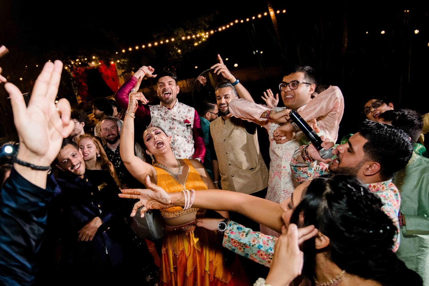 Energetic crowd dancing and celebrating at a lively wedding reception with string lights overhead.