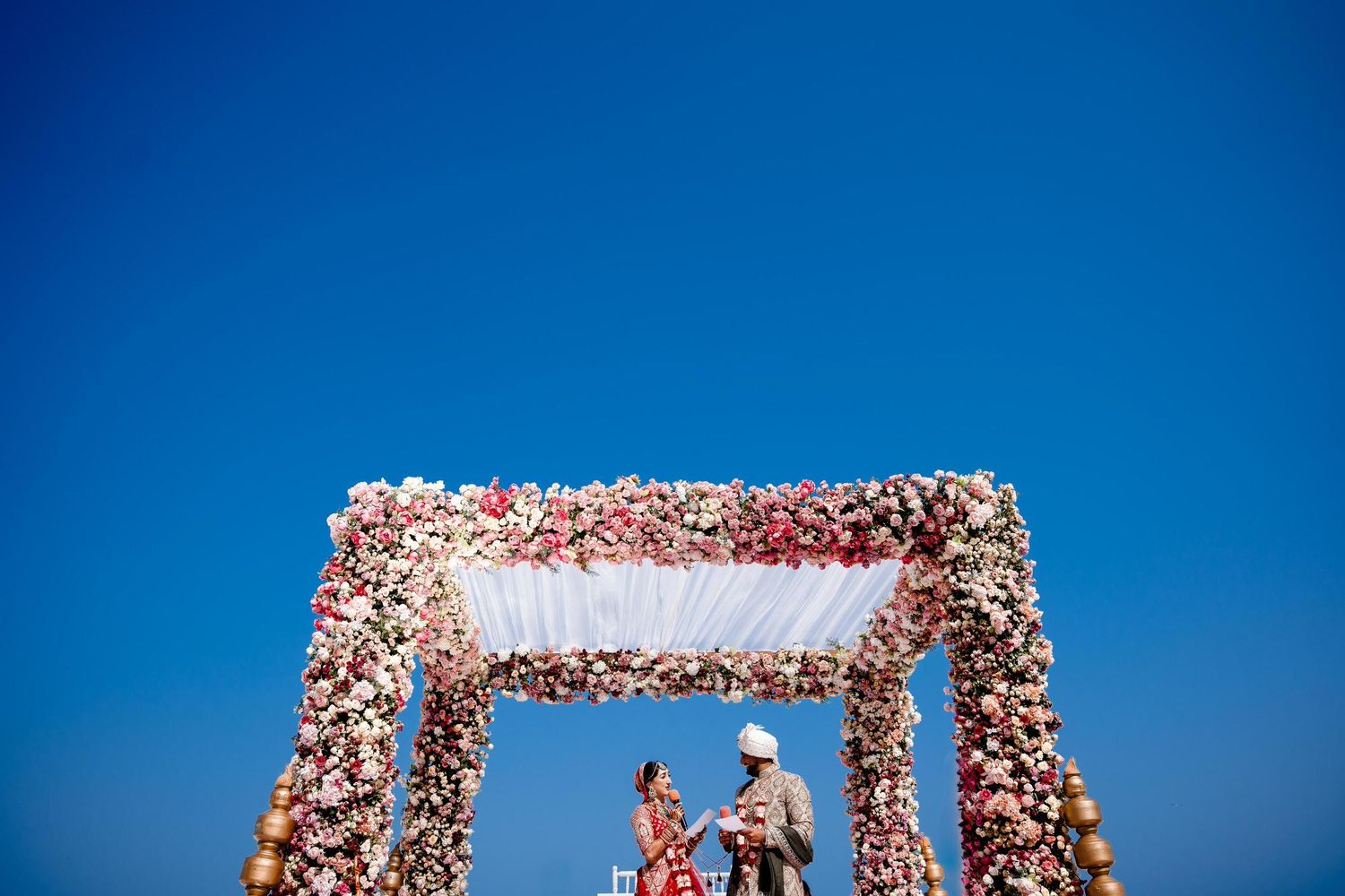 A floral wedding arch with pink and white flowers stands against a bright blue sky during an outdoor ceremony.