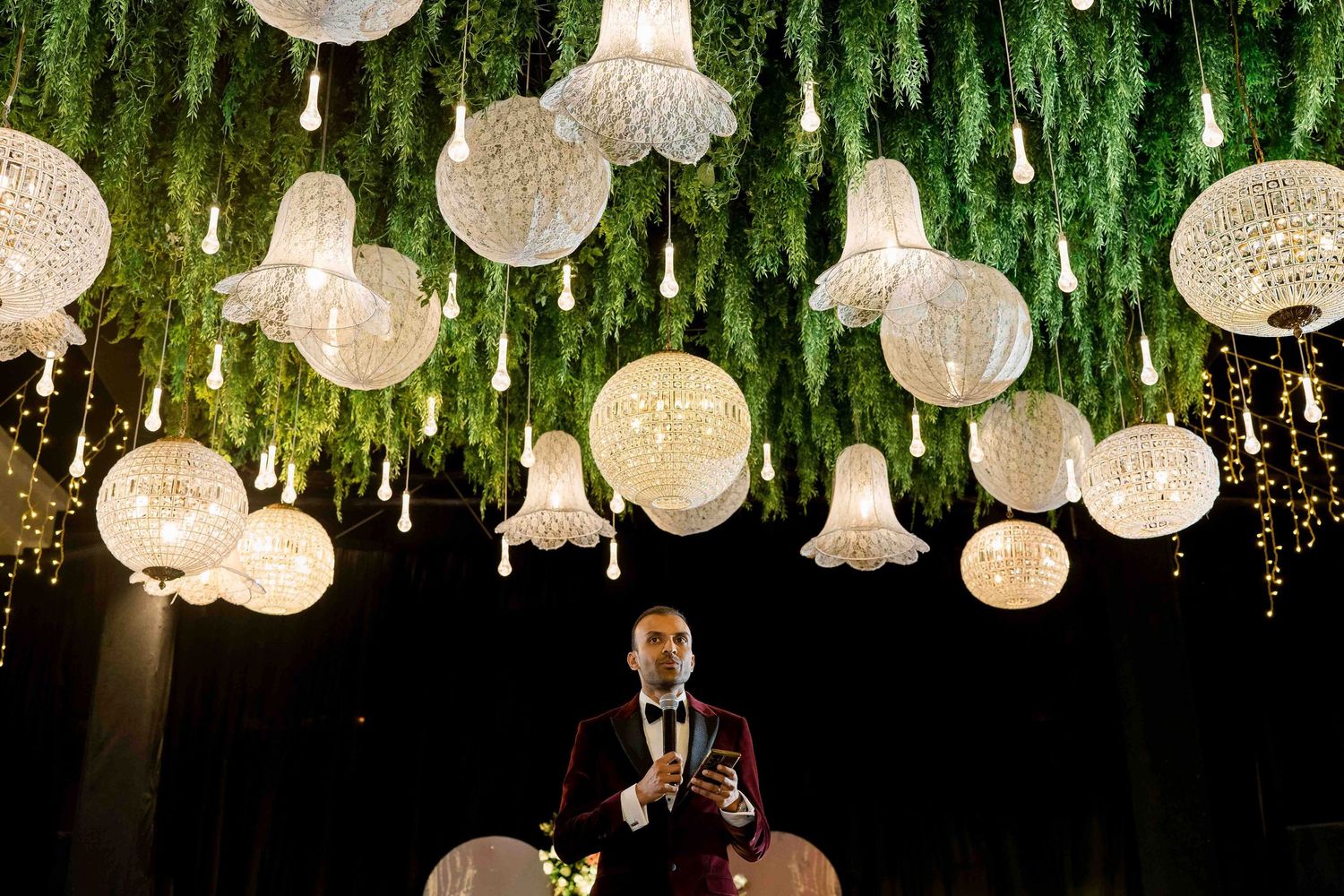 Crystal chandeliers and orb-shaped pendant lights hang elegantly from a dark ceiling with cascading greenery decorations.