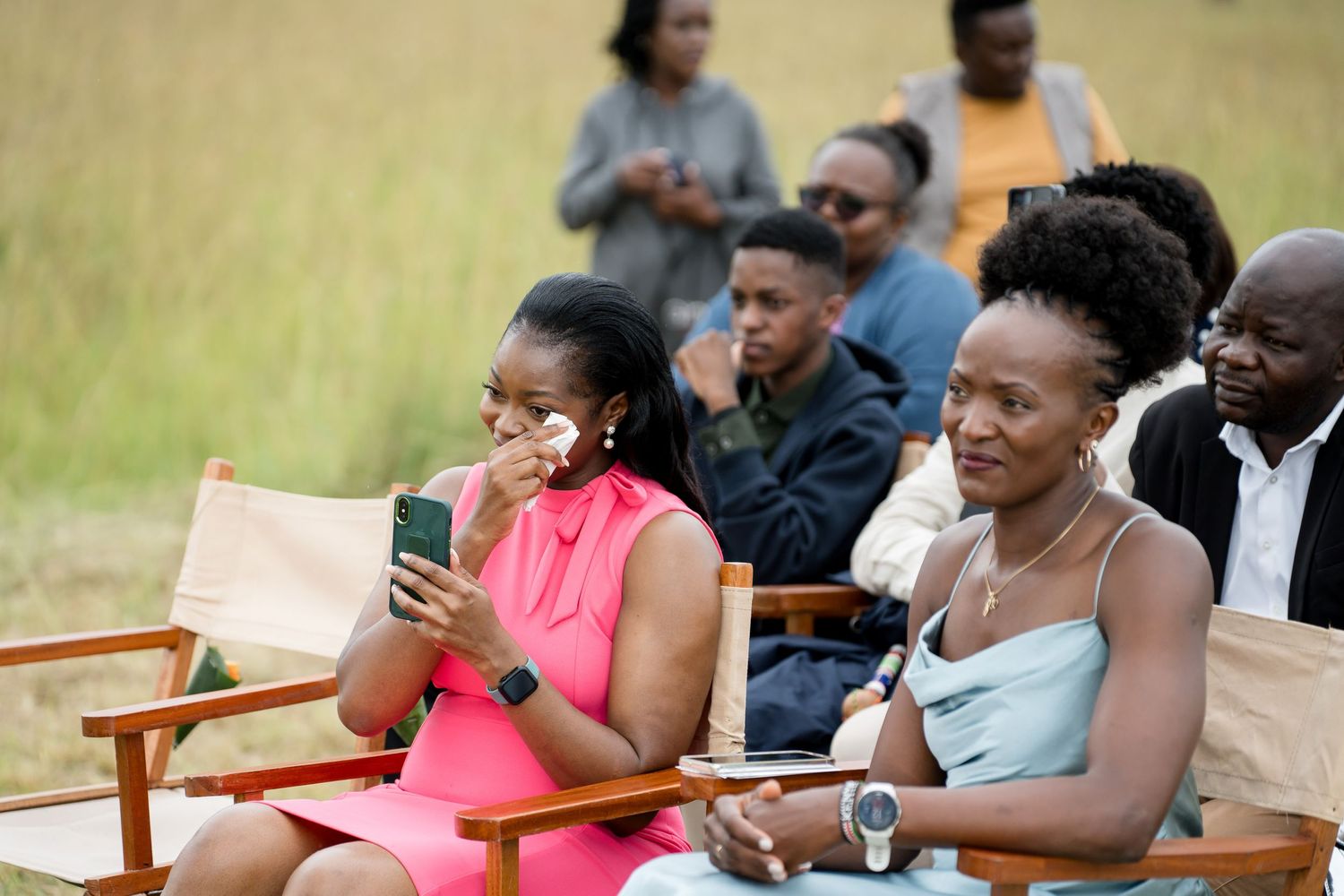 Wedding guests in bright attire watch ceremony intently from wooden chairs.