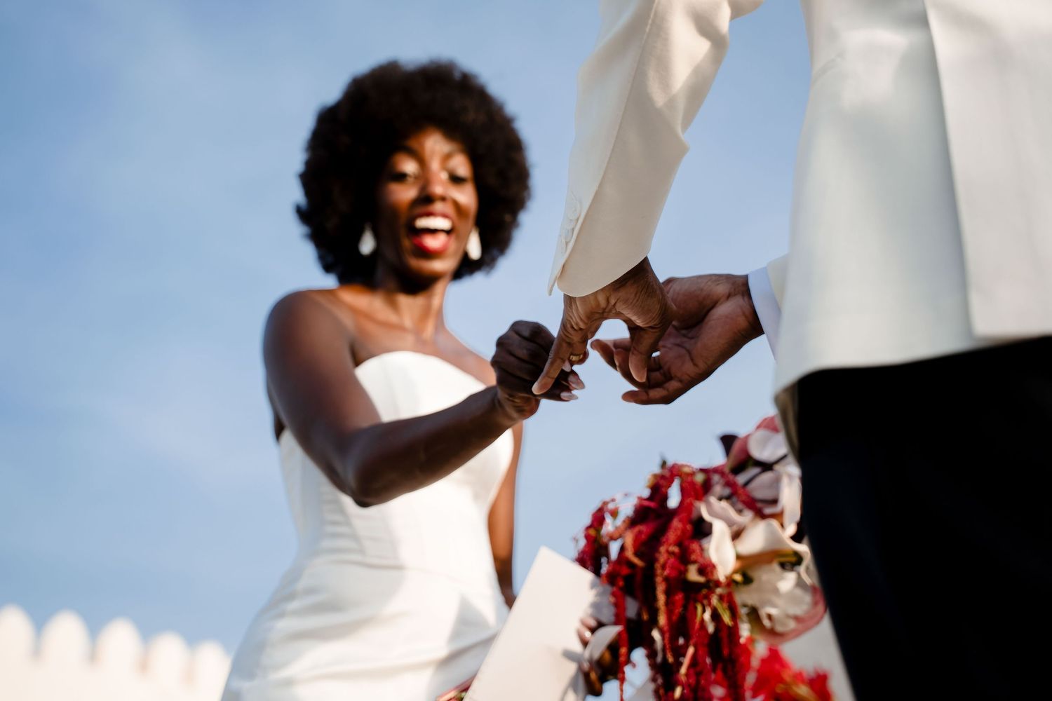 A joyful bride in a white wedding dress holds a vibrant red bouquet while dancing and celebrating.
