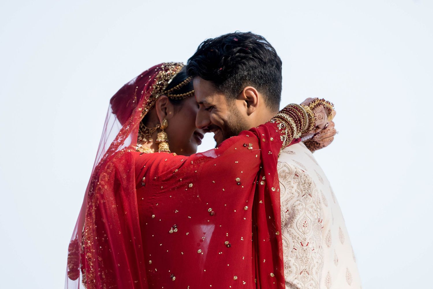 Two people in traditional wedding attire embrace under a red veil during a romantic ceremony moment.