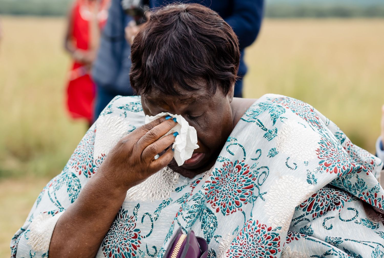 Wedding guest with floral print blanket dabs tears with tissue during emotional outdoor ceremony.