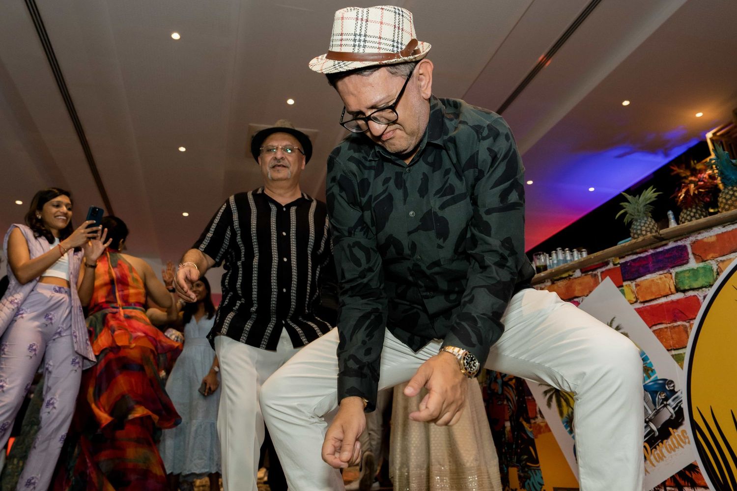 People dressed in retro outfits dance enthusiastically during a party with colorful lighting and decorations in the background.