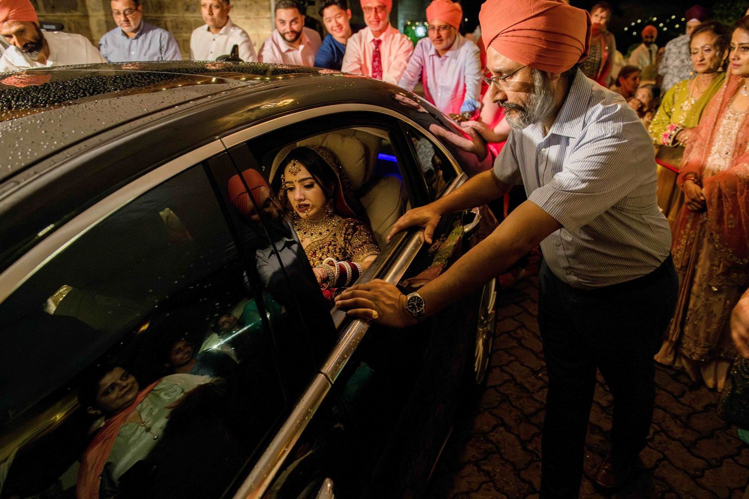 A crowd gathers around a car during a night celebration as passengers prepare to depart.