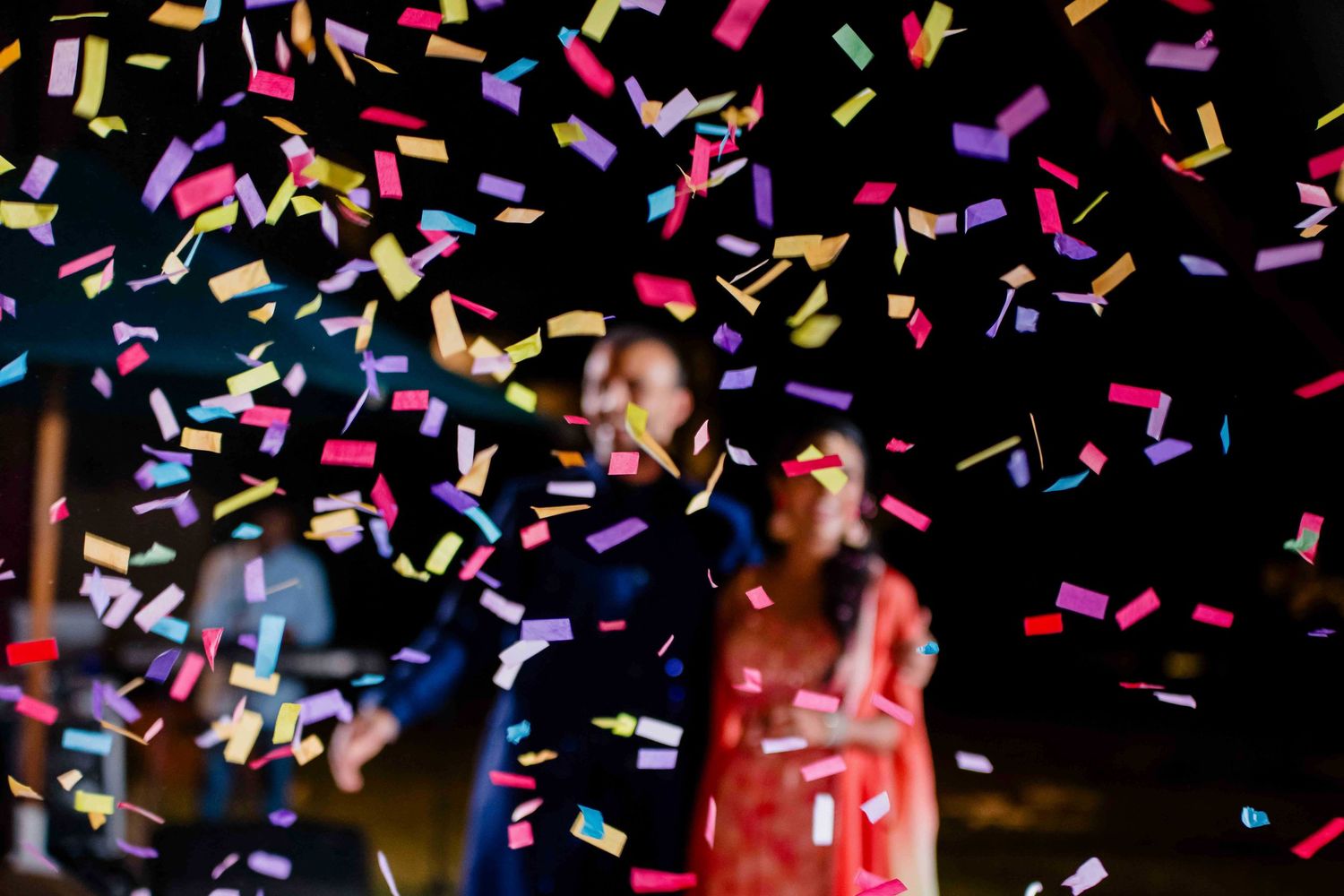 Colorful confetti falls against a dark background during a celebration with blurred stage lights and figures.