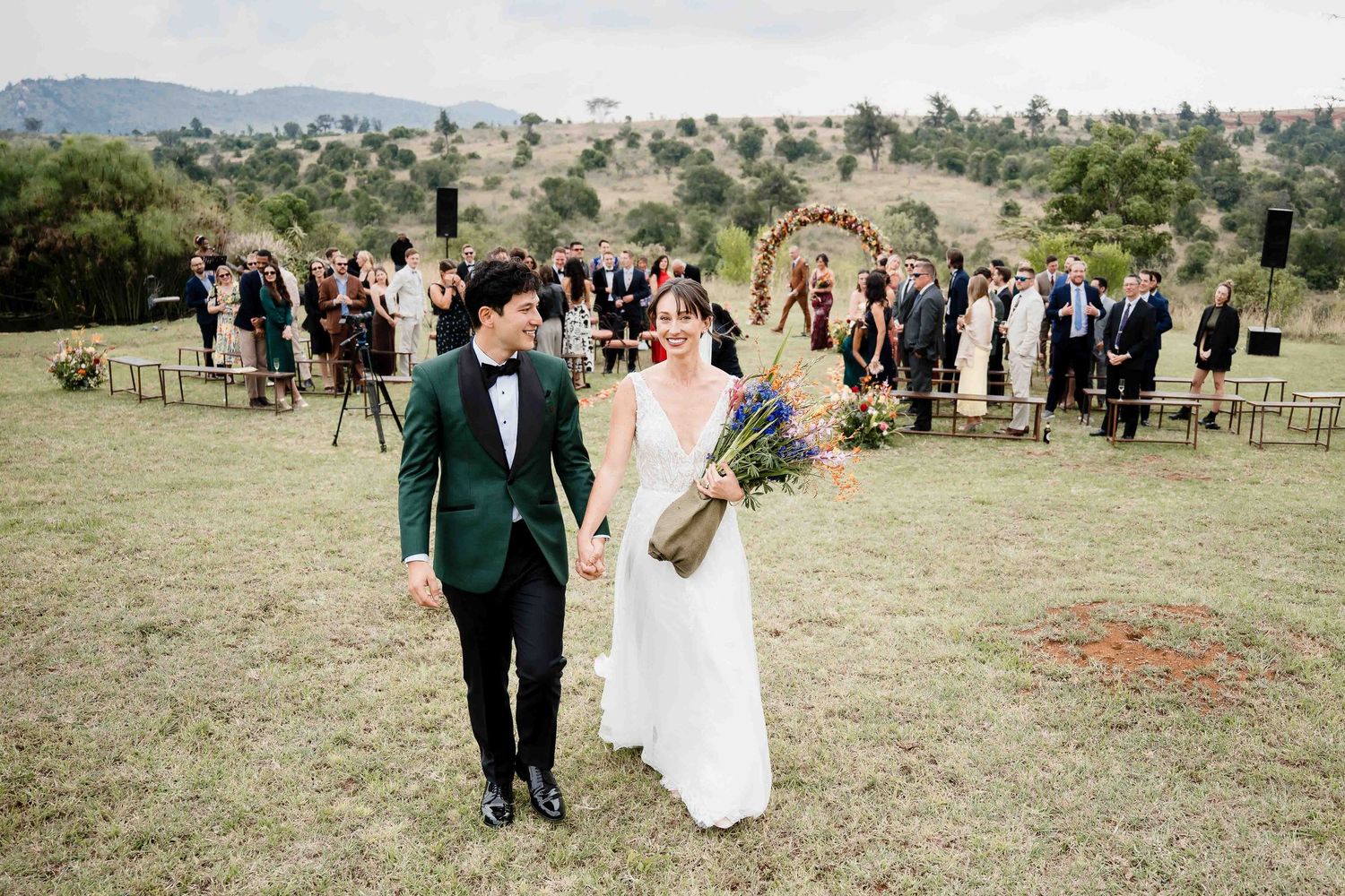 An outdoor wedding ceremony on a hilltop with guests gathered behind the couple in formal attire.