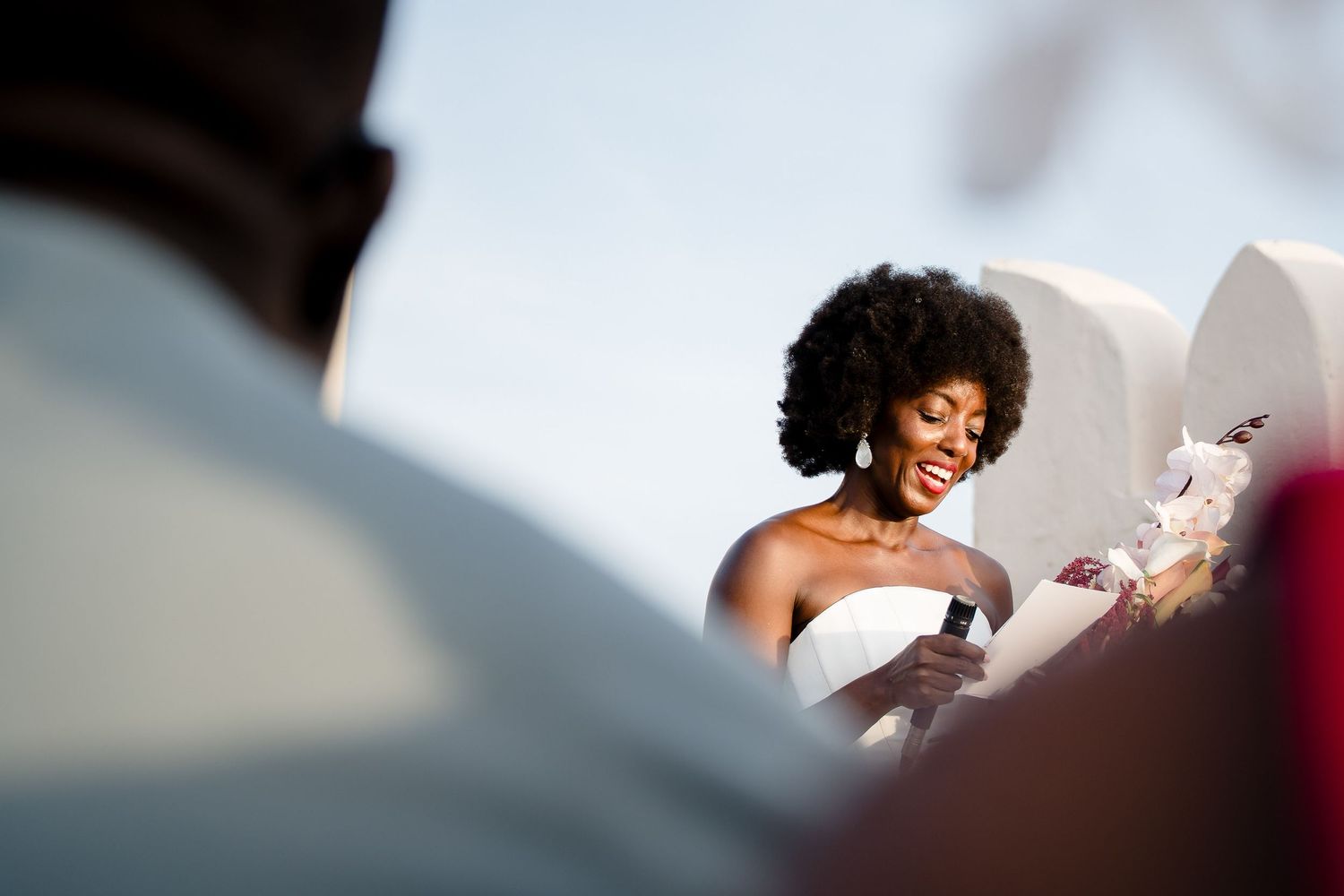 A bride in a white dress with natural curly hair smiles while reading from papers during a wedding ceremony.