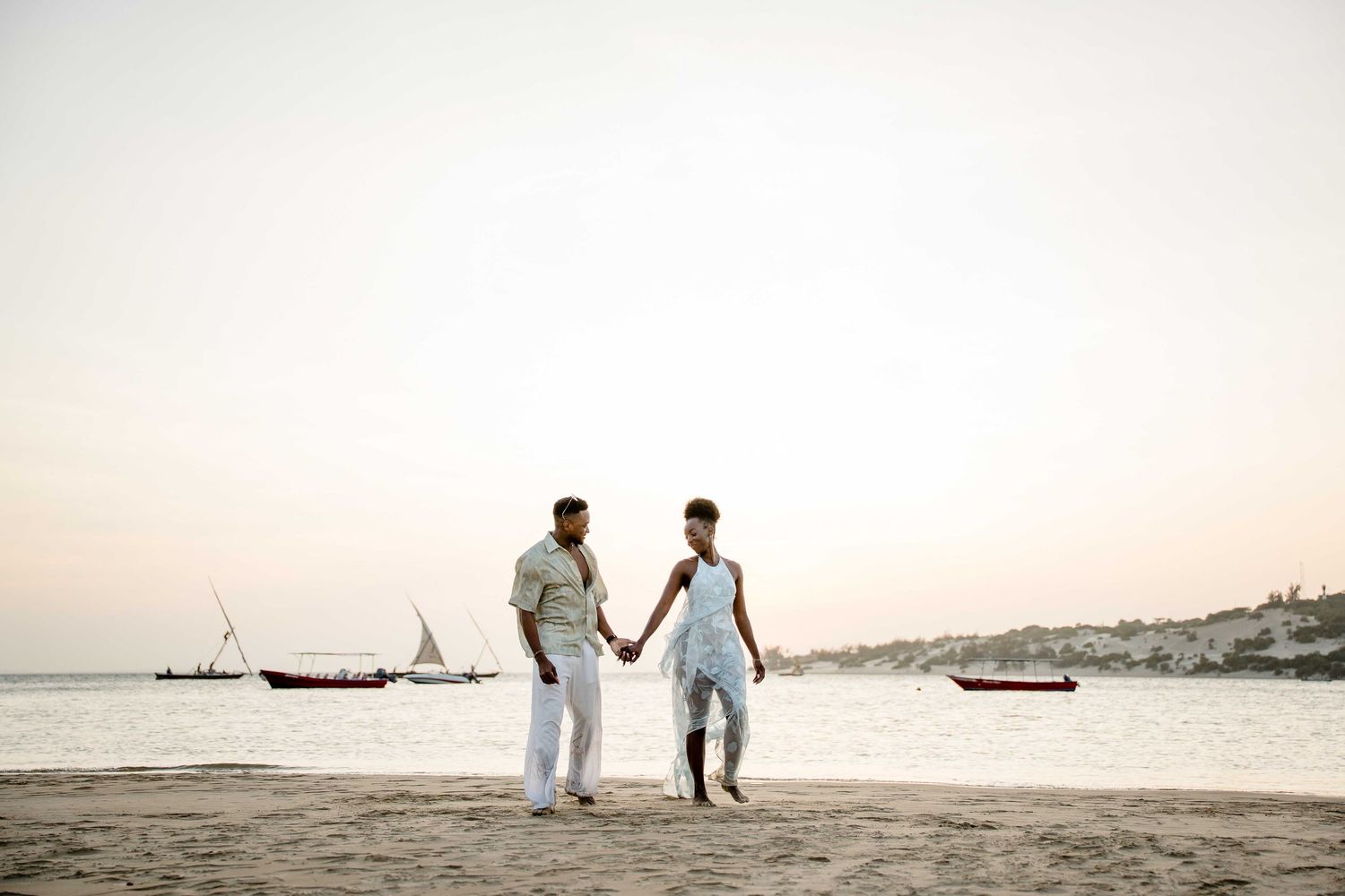 Two people in white outfits walk along a scenic beach at sunset with boats visible in the background.